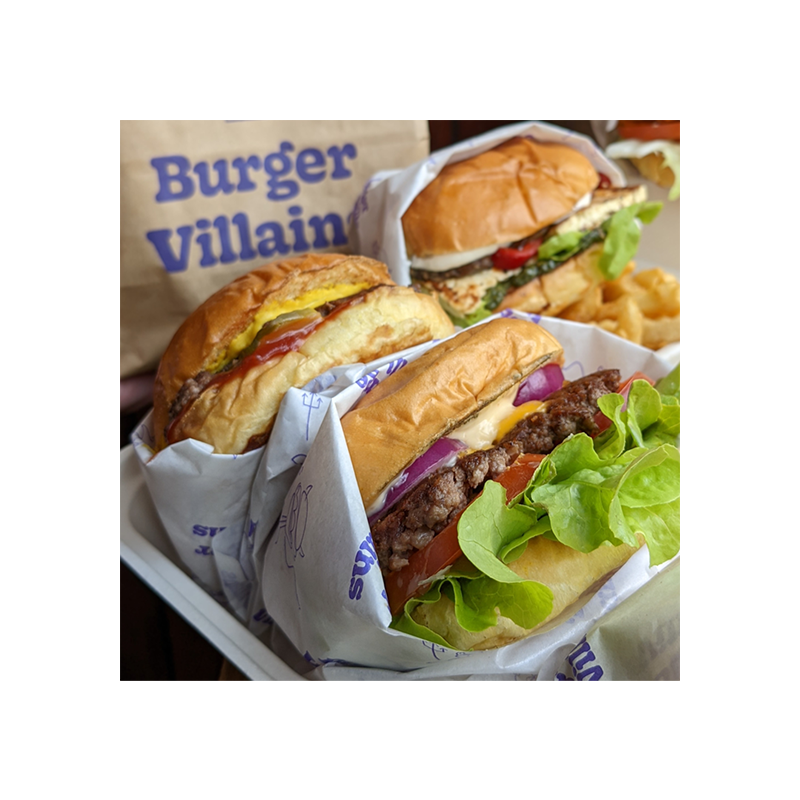 Three Burger Villains burgers wrapped in the store's packaging, in front of a brown bag with the purple Burger Villains logo