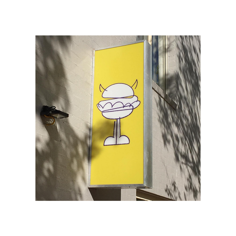 A yellow vertical sign, with the Burger Villains mascot logo on it. The sign is hanging above the Burger Villains store in Phillip